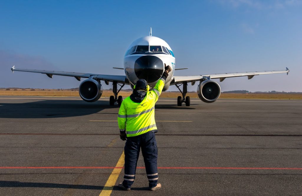 An airport worker in a high-visibility jacket guides a large commercial airplane on the runway.